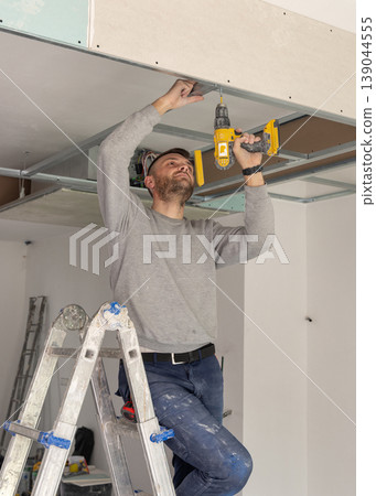 Construction worker using drill on metal frame for drywall suspended ceiling in apartment renovation Construction worker using drill on metal frame for drywall suspended ceiling in apartment renovation 139044555