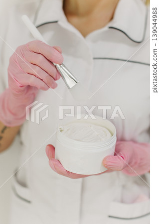 Facial clay mask in white jar and brush in a hands of a cosmetologist. Skin care beauty product. Cosmetology concept. 139044988