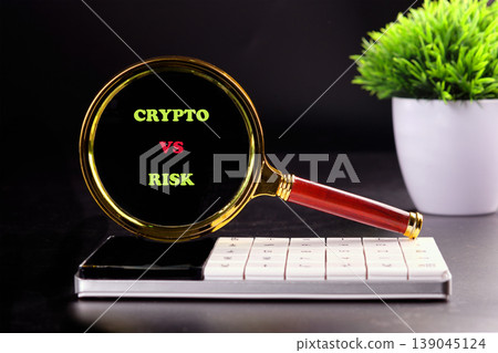 A magnifying glass displays Crypto vs Risk over a white calculator and phone on a dark surface, near a green potted plant A magnifying glass displays Crypto vs Risk over a white calculator and phone on a dark surface, near a green potted plant 139045124