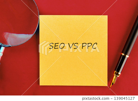 A yellow sticky note displays SEO VS PPC in black text. A magnifying glass and fountain pen are arranged on a vibrant red surface A yellow sticky note displays SEO VS PPC in black text. A magnifying glass and fountain pen are arranged on a vibrant red surface 139045127