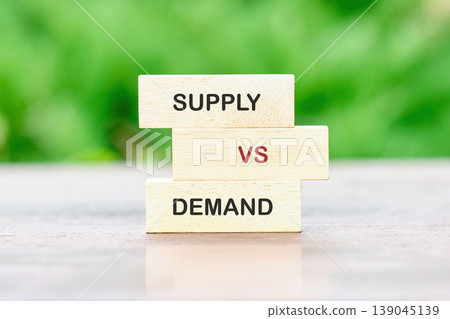 Three wooden blocks stacked vertically on a light wooden table. The top block reads SUPPLY, the middle VS in red, and the bottom DEMAND Three wooden blocks stacked vertically on a light wooden table. The top block reads SUPPLY, the middle VS in red, and the bottom DEMAND 139045139