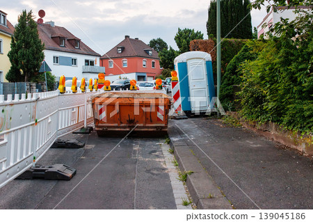 Orange construction waste container with amber warning lights and red white striped barriers, with a temporary blue portable toilet booth on the roadside Orange construction waste container with amber warning lights and red white striped barriers, with a temporary blue portable toilet booth on the roadside 139045186