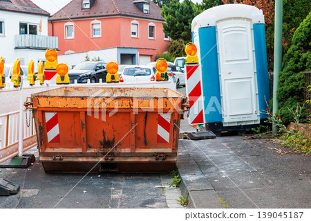 Orange construction waste container with amber warning lights and red white striped barriers, with a temporary blue portable toilet booth on the roadside Orange construction waste container with amber warning lights and red white striped barriers, with a temporary blue portable toilet booth on the roadside 139045187