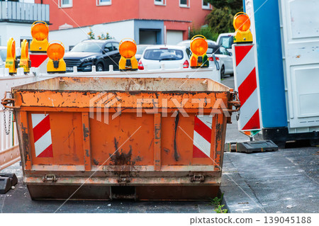 Orange construction waste container with amber warning lights and red white striped barriers, with a temporary blue portable toilet booth on the roadside Orange construction waste container with amber warning lights and red white striped barriers, with a temporary blue portable toilet booth on the roadside 139045188