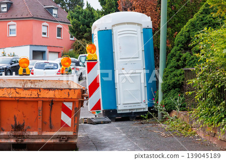 Orange construction waste container with amber warning lights and red white striped barriers, with a temporary blue portable toilet booth on the roadside 139045189