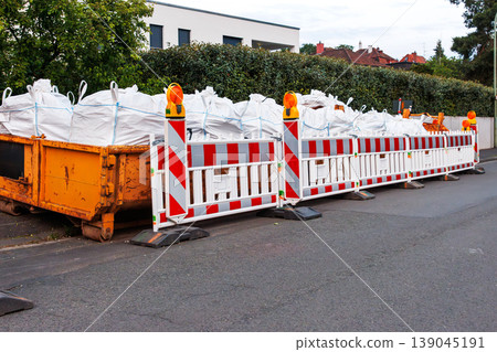 Urban roadwork area with red and white barriers and orange warning beacons, with white bulk sacks of construction materials waste in waste container 139045191
