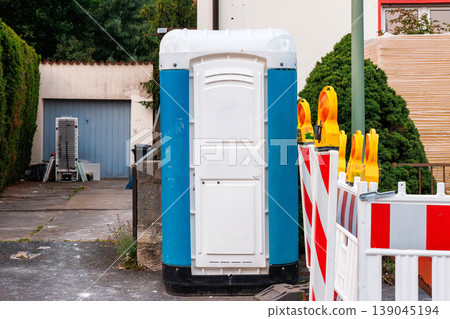 Portable toilet booth beside roadwork barriers with warning beacons. Discarded old fridge and household items in driveway before waste pickup Portable toilet booth beside roadwork barriers with warning beacons. Discarded old fridge and household items in driveway before waste pickup 139045194