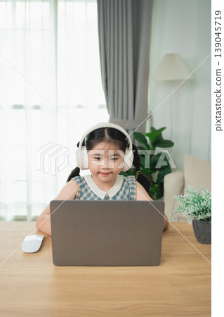 Young girl wearing headphones sitting at a desk with a laptop in a cozy living room, focused on online learning or entertainment in a bright environment 139045719