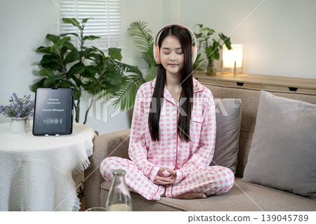 Asian young adult woman meditating relaxing home environment using headphones listening calming music for stress relief mindfulness practice Asian young adult woman meditating relaxing home environment using headphones listening calming music for stress relief mindfulness practice 139045789