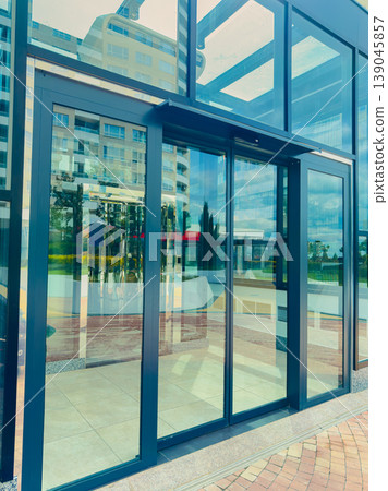Modern glass entrance of a building showing reflection and surroundings in the afternoon light with clear skies and urban setting Modern glass entrance of a building showing reflection and surroundings in the afternoon light with clear skies and urban setting 139045857