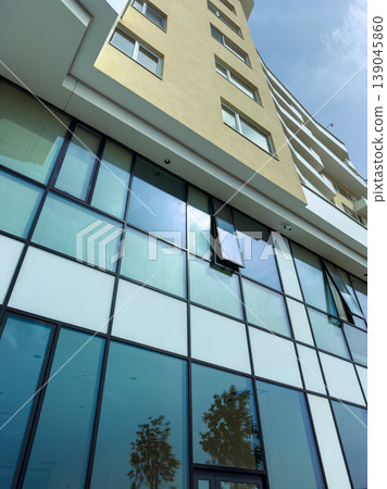 Looking up at a modern building with glass windows and a clear sky during the day in an urban area with trees nearby Looking up at a modern building with glass windows and a clear sky during the day in an urban area with trees nearby 139045860