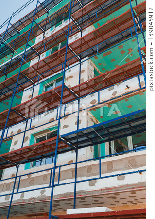 Construction work is in progress on a building with scaffolding and green sheeting covering parts of its exterior in an urban area during daylight hours Construction work is in progress on a building with scaffolding and green sheeting covering parts of its exterior in an urban area during daylight hours 139045912