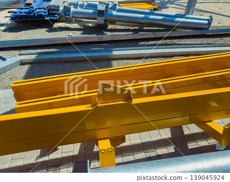 Construction site shows yellow beams and metal pipes arranged for assembly during daylight, with tools and materials nearby indicating ongoing work 139045924