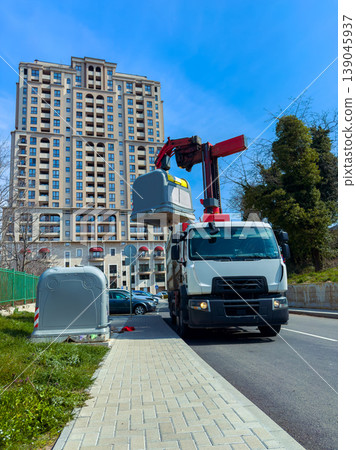 Garbage truck lifts bins for collection in urban area with high-rise building in background during daytime Garbage truck lifts bins for collection in urban area with high-rise building in background during daytime 139045937