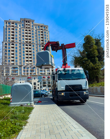Truck lifts garbage bin on street in urban area near tall building under clear blue sky during daylight hours Truck lifts garbage bin on street in urban area near tall building under clear blue sky during daylight hours 139045938
