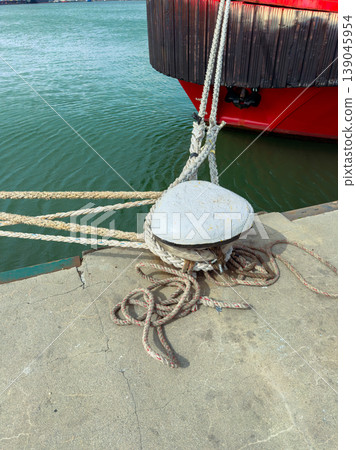 Rope tied to a mooring cleat at a busy dock during the afternoon with a boat anchored nearby 139045954
