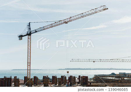 Construction work takes place by the water's edge with cranes, buildings, and workers under a clear sky during the day in an urban area 139046085