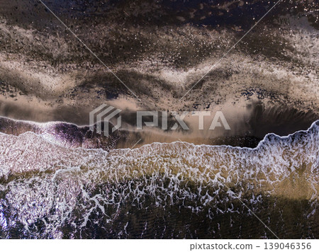 Waves crash against the shore during sunset at a beach, captured from above showing patterns in water and sand Waves crash against the shore during sunset at a beach, captured from above showing patterns in water and sand 139046356