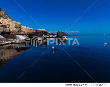 Bright blue sky above calm water with rocks and seagulls near the shore at midday Bright blue sky above calm water with rocks and seagulls near the shore at midday 139046535