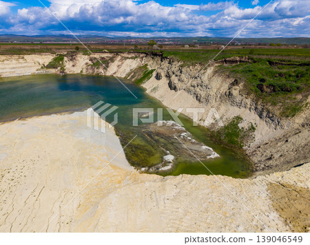 Clouds float above a water-filled pit in a landscape with grass, sand, and rocky edges in a rural area during daytime Clouds float above a water-filled pit in a landscape with grass, sand, and rocky edges in a rural area during daytime 139046549