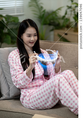 Asian young adult woman uses LED light therapy device for facial skin rejuvenation in pajamas home setting 139047046