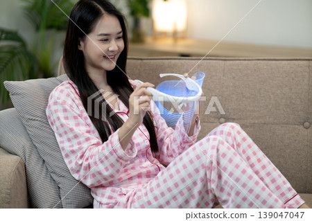 Asian young adult woman home beauty device using light therapy skin care facial treatment relaxing comfortable sofa living room self care routine wellness technology enjoyment Asian young adult woman home beauty device using light therapy skin care facial treatment relaxing comfortable sofa living room self care routine wellness technology enjoyment 139047047