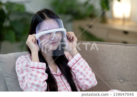 Asian adult woman LED light therapy mask skincare beauty home couch relax living room facial wellness Asian adult woman LED light therapy mask skincare beauty home couch relax living room facial wellness 139047048