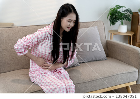 Asian adult woman feeling severe stomach pain abdominal cramps discomfort unwell at home couch indoor wearing pajamas suffering menstrual health issues digestive sickness Asian adult woman feeling severe stomach pain abdominal cramps discomfort unwell at home couch indoor wearing pajamas suffering menstrual health issues digestive sickness 139047051