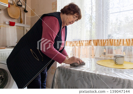 Senior Caucasian woman cleaning the kitchen table alone at home. Daily routine and hard household chores in retirement. Concept of loneliness, aging, and social isolation 139047144