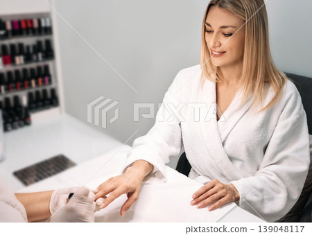 Beautiful woman in bathrobe during manicure in spa salon, taking care of her nails. Manicurist covers woman's nails with nail polish Beautiful woman in bathrobe during manicure in spa salon, taking care of her nails. Manicurist covers woman's nails with nail polish 139048117