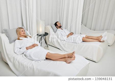 Couple while romantic weekend. Loving young couple in white bathrobes spending time at spa laying down on white beds in lounge zone Couple while romantic weekend. Loving young couple in white bathrobes spending time at spa laying down on white beds in lounge zone 139048131