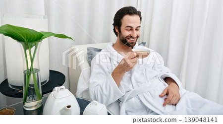 Portrait of handsome man in white bathrobe drinking healing tea while relaxing in wellness center. Spa rest for men Portrait of handsome man in white bathrobe drinking healing tea while relaxing in wellness center. Spa rest for men 139048141