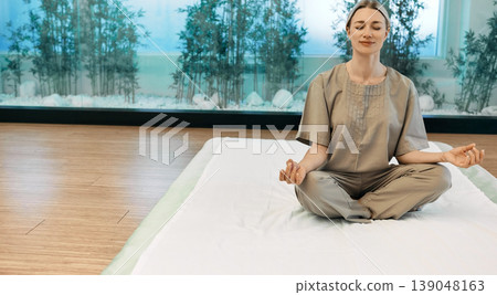 Attractive woman sitting in lotus pose or asana Padmasana on yoga mat with closed eyes in meditation room with bamboo. Yoga, meditation, breathing exercises Attractive woman sitting in lotus pose or asana Padmasana on yoga mat with closed eyes in meditation room with bamboo. Yoga, meditation, breathing exercises 139048163