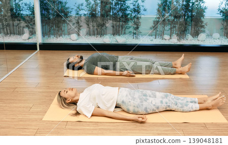 Fitness couple in sportswear practice yoga while lying down in savasana or corpse pose at yoga center. Shavasana is important pose in restorative yoga Fitness couple in sportswear practice yoga while lying down in savasana or corpse pose at yoga center. Shavasana is important pose in restorative yoga 139048181