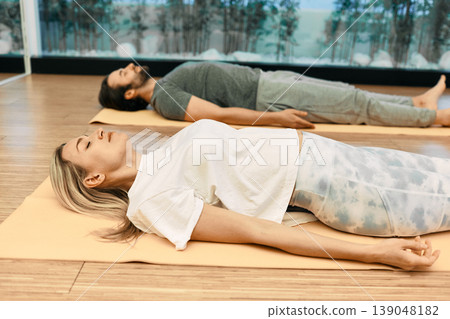 Shavasana. Man with woman wearing in sportswear practice yoga while lying down in savasana or corpse pose at wellness center Shavasana. Man with woman wearing in sportswear practice yoga while lying down in savasana or corpse pose at wellness center 139048182