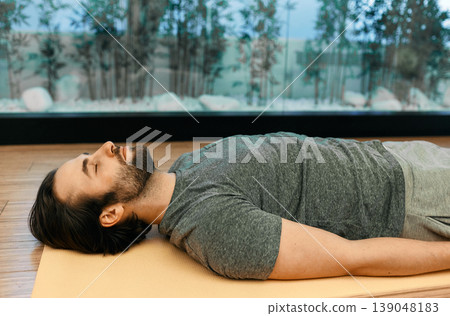Savasana or corpse dead pose. Handsome man relaxes in motionless corpse pose with closed eyes lying on floor of meditation center Savasana or corpse dead pose. Handsome man relaxes in motionless corpse pose with closed eyes lying on floor of meditation center 139048183