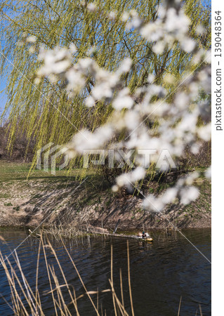 Ducks relaxing in a pond, a spring willow, and blooming cherry plums over a canal, a vertical poster about spring and a weekend getaway in nature 139048364