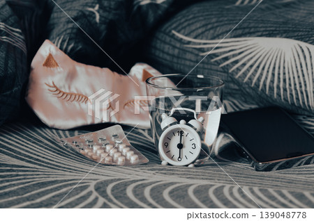 Sleeping pills, a glass of water, a mask, and an alarm clock on the bed Sleeping pills, a glass of water, a mask, and an alarm clock on the bed 139048778