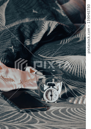 Sleeping pills, a glass of water, a mask, and an alarm clock on the bed vertical view Sleeping pills, a glass of water, a mask, and an alarm clock on the bed vertical view 139048780