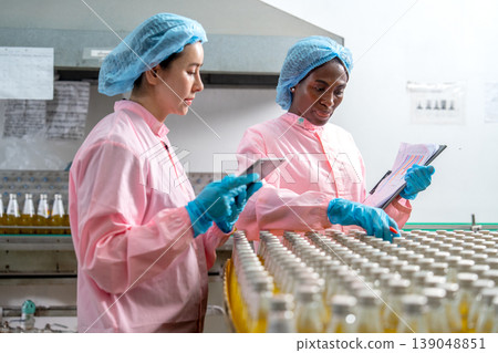 Side view of Asian and African American factory woman workers stand and check quality in the bottle in front of the containing machine in the factory workplace. 139048851