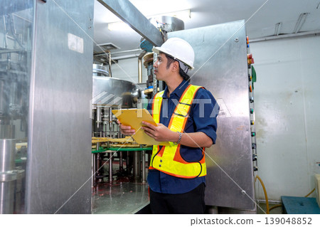 Side view and wide shot of Asian factory technician or engineer hold document file and stand in front of containing machine to check the quality of the line product and function of machine with 139048852