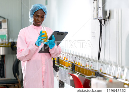 African American woman factory worker who hold document file and bottle with yellow liquid to check quality of product in line productivity in drinking industrial factory workplace. African American woman factory worker who hold document file and bottle with yellow liquid to check quality of product in line productivity in drinking industrial factory workplace. 139048862