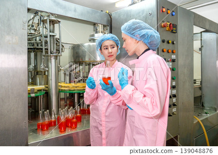 Side view of two factory woman workers discuss about the quality and smell of red drinking liquid in the bottle in front of the containing machine in the factory workplace. 139048876