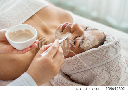 Esthetician applying white clay face mask with spatula on young black woman 139049802