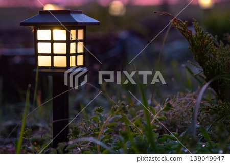 Modern garden design with solar powered lantern, mixed ornamental grasses and perennials, mulched bed, sustainable outdoor lighting, evening ambiance. 139049947