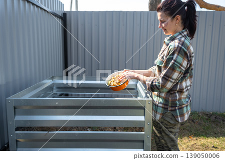 Adding kitchen food scraps and grass to metal compost bin, home composting process, sustainable organic waste reuse in steel frame structure. Adding kitchen food scraps and grass to metal compost bin, home composting process, sustainable organic waste reuse in steel frame structure. 139050006