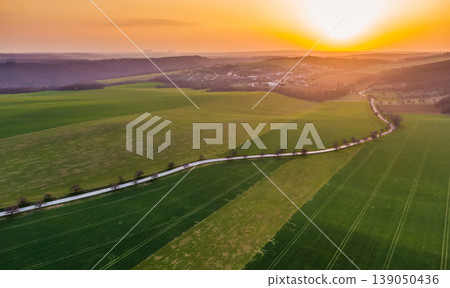 Aerial view of agriculture land during beautiful sunset. Fields and meadow with the small village in far. Vibrant soft colors at magic dusk. The asphalt road leading between fields with tree on side Aerial view of agriculture land during beautiful sunset. Fields and meadow with the small village in far. Vibrant soft colors at magic dusk. The asphalt road leading between fields with tree on side 139050436