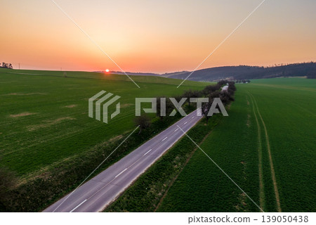 Aerial view of agriculture land during beautiful sunset. Fields and meadow with the small village in far. Vibrant soft colors at magic dusk. The asphalt road leading between fields with tree on side Aerial view of agriculture land during beautiful sunset. Fields and meadow with the small village in far. Vibrant soft colors at magic dusk. The asphalt road leading between fields with tree on side 139050438