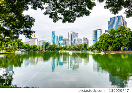 Bangkok cityscape view from Lumpini park in city center. Panoramic photo of skyscrapers and futuristic buildings from metal and glass. View from the small lake in Lumpini park. Fresh green plants. Bangkok cityscape view from Lumpini park in city center. Panoramic photo of skyscrapers and futuristic buildings from metal and glass. View from the small lake in Lumpini park. Fresh green plants. 139050747
