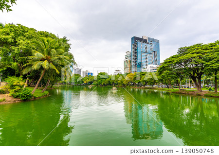 Bangkok cityscape view from Lumpini park in city center. Panoramic photo of skyscrapers and futuristic buildings from metal and glass. View from the small lake in Lumpini park. Fresh green plants. Bangkok cityscape view from Lumpini park in city center. Panoramic photo of skyscrapers and futuristic buildings from metal and glass. View from the small lake in Lumpini park. Fresh green plants. 139050748